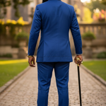 Man in a blue Aso Oke suit walking with a cane on a cobblestone path.
