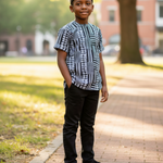 Child wearing a black white Kente dashiki shirt and black pants standing on a sidewalk with trees and buildings in the background.